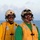 Two military personnel are standing on what appears to be a naval vessel deck with open ocean visible in the background. Both individuals are wearing yellow safety vests, protective goggles, helmets with communication headsets, and microphones, indicating they are engaged in flight deck or maritime operations. The image was likely taken during dawn or dusk based on the soft lighting conditions.