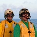 Two military personnel are standing on what appears to be a naval vessel deck with open ocean visible in the background. Both individuals are wearing yellow safety vests, protective goggles, helmets with communication headsets, and microphones, indicating they are engaged in flight deck or maritime operations. The image was likely taken during dawn or dusk based on the soft lighting conditions.