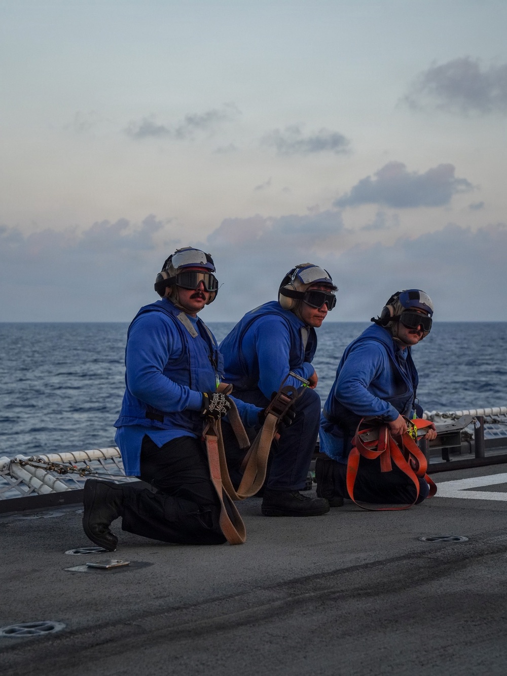 Three naval personnel in blue flight deck jerseys are kneeling on what appears to be a ship's flight deck or helicopter landing pad at dusk or dawn. They are equipped with cranial helmets, ear protection, and goggles, indicating active flight deck operations. Each person is holding or positioned near tie-down straps and equipment bags used for securing aircraft.