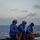 Three naval personnel in blue flight deck jerseys are kneeling on what appears to be a ship's flight deck or helicopter landing pad at dusk or dawn. They are equipped with cranial helmets, ear protection, and goggles, indicating active flight deck operations. Each person is holding or positioned near tie-down straps and equipment bags used for securing aircraft.