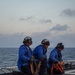 Three naval personnel in blue flight deck jerseys are kneeling on what appears to be a ship's flight deck or helicopter landing pad at dusk or dawn. They are equipped with cranial helmets, ear protection, and goggles, indicating active flight deck operations. Each person is holding or positioned near tie-down straps and equipment bags used for securing aircraft.
