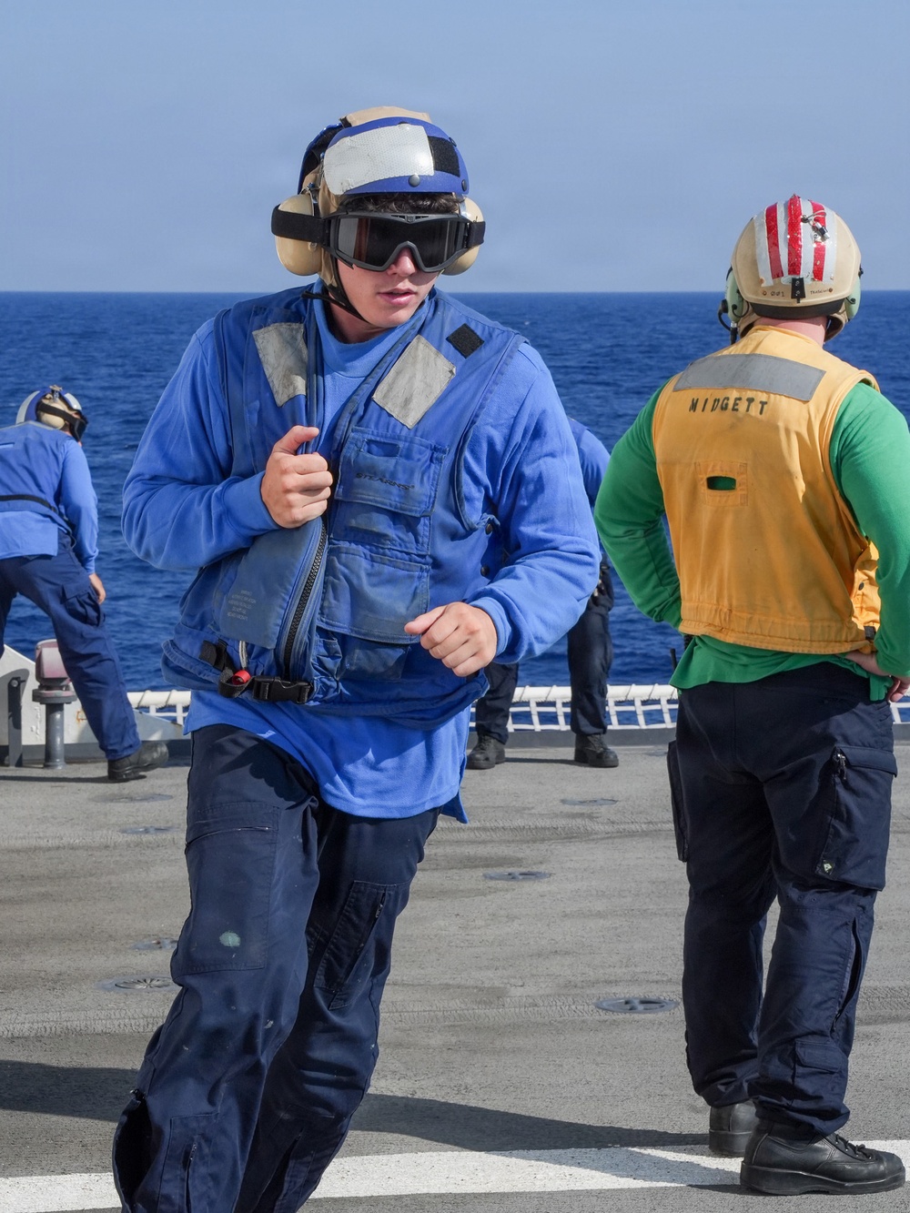 The image shows U.S. Navy personnel on the flight deck of a naval vessel at sea. The foreground sailor is wearing blue coveralls, a blue float coat/life preserver, a blue helmet with goggles, and hearing protection ear muffs. A second sailor in the background wears a yellow vest labeled 'WIDGETT' and a red-striped helmet, indicating a specific flight deck role.
