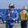 The image shows U.S. Navy personnel on the flight deck of a naval vessel at sea. The foreground sailor is wearing blue coveralls, a blue float coat/life preserver, a blue helmet with goggles, and hearing protection ear muffs. A second sailor in the background wears a yellow vest labeled 'WIDGETT' and a red-striped helmet, indicating a specific flight deck role.