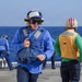 The image shows U.S. Navy personnel on the flight deck of a naval vessel at sea. The foreground sailor is wearing blue coveralls, a blue float coat/life preserver, a blue helmet with goggles, and hearing protection ear muffs. A second sailor in the background wears a yellow vest labeled 'WIDGETT' and a red-striped helmet, indicating a specific flight deck role.