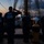 Four individuals stand on the wooden deck of a tall ship at sunset or sunrise, with one person rendering a salute. They are wearing matching navy blue t-shirts reading 'Barque Eagle - America's Tall Ship' and at least one shirt reading 'SAIL Summer 2020'. The scene is backlit by warm orange and purple twilight sky over calm water.