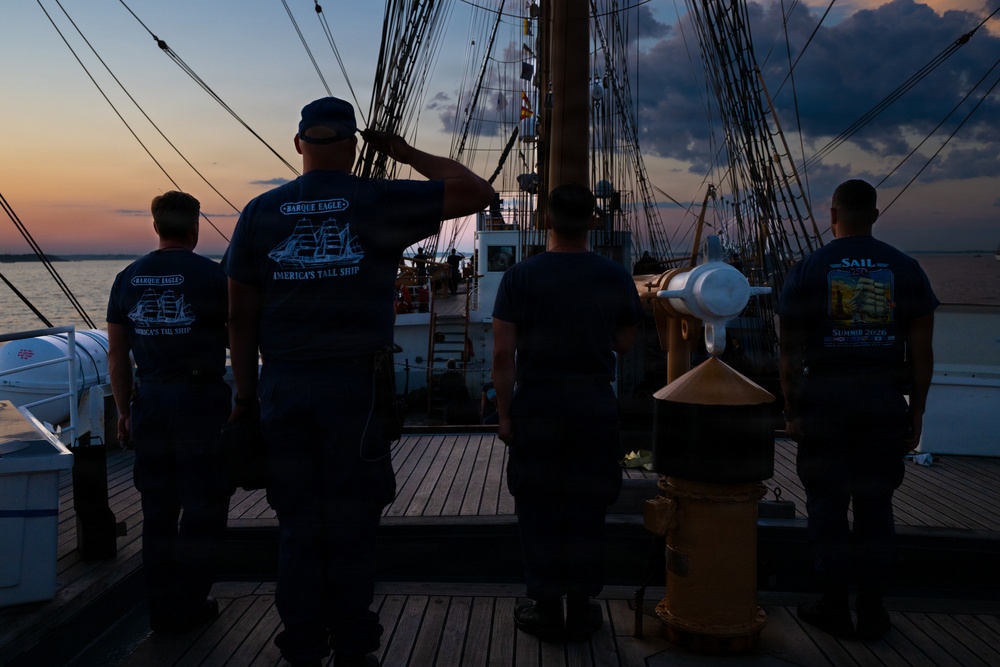 Four individuals stand on the wooden deck of a tall ship at sunset or sunrise, with at least one person rendering a salute. They are wearing matching navy blue t-shirts reading 'Barque Eagle - America's Tall Ship' and one shirt reading 'Sail Summer 2020'. The scene is backlit by warm orange and purple twilight sky over calm water.