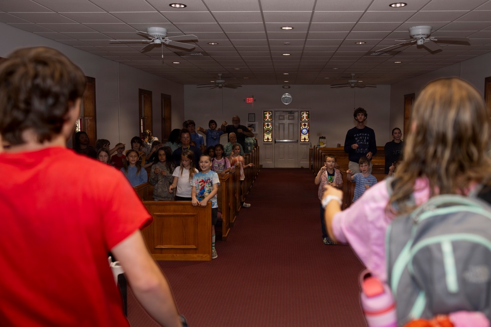 A group of children and adults are gathered inside a church sanctuary with wooden pews and red carpet. The scene appears to be an interactive activity or game, with children standing in the aisle and between pews in animated poses. Two individuals in the foreground (viewed from behind) appear to be leading or facilitating the activity.