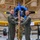 A military change of command ceremony is taking place inside what appears to be an aviation museum or hangar. Two personnel in olive green flight suits are jointly holding a blue guidon (unit flag) bearing the number '28', while a third service member in OCP camouflage uniform stands behind them holding the flagstaff. A vintage WWII-era training aircraft with a blue engine cowling and yellow wings is prominently displayed in the background.