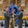 A military change of command ceremony taking place inside what appears to be an aviation museum or hangar. Two officers in olive green flight suits are jointly holding a unit guidon (flag on a staff), with a third service member in camouflage uniform standing behind them. A vintage yellow and blue propeller aircraft is prominently displayed in the background.