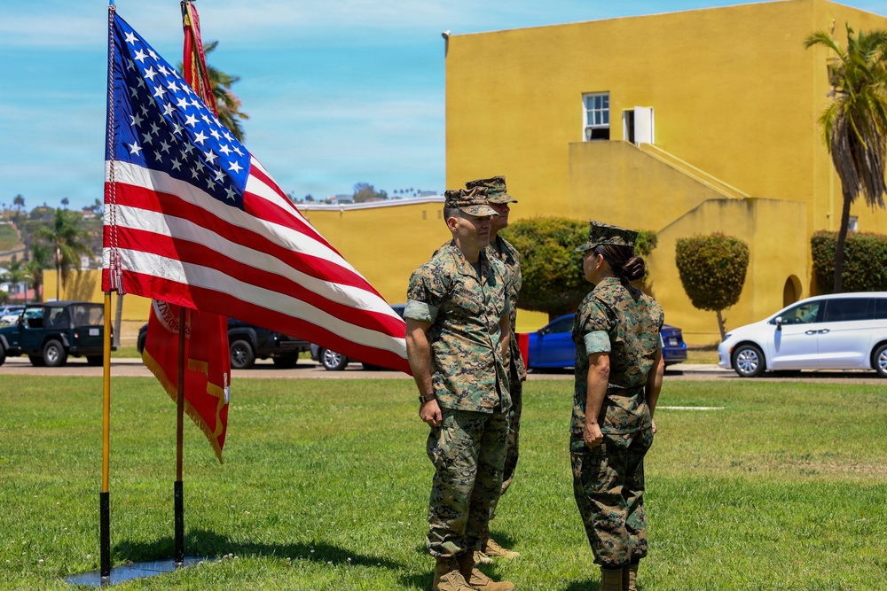 Two U.S. Marine Corps personnel in MARPAT woodland camouflage uniforms stand on a grass parade field next to a color guard display featuring the American flag and what appears to be a Marine Corps unit guidon. The setting appears to be a military base, likely in Southern California based on the architecture, palm trees, and yellow stucco building visible in the background. The scene suggests a formal military ceremony such as a change of command, promotion, or unit formation.