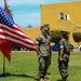 Two U.S. Marine Corps personnel in MARPAT woodland camouflage uniforms stand on a grass parade field next to a color guard display featuring the American flag and what appears to be a Marine Corps unit guidon. The setting appears to be a military base, likely in Southern California based on the architecture, palm trees, and yellow stucco building visible in the background. The scene suggests a formal military ceremony such as a change of command, promotion, or unit formation.