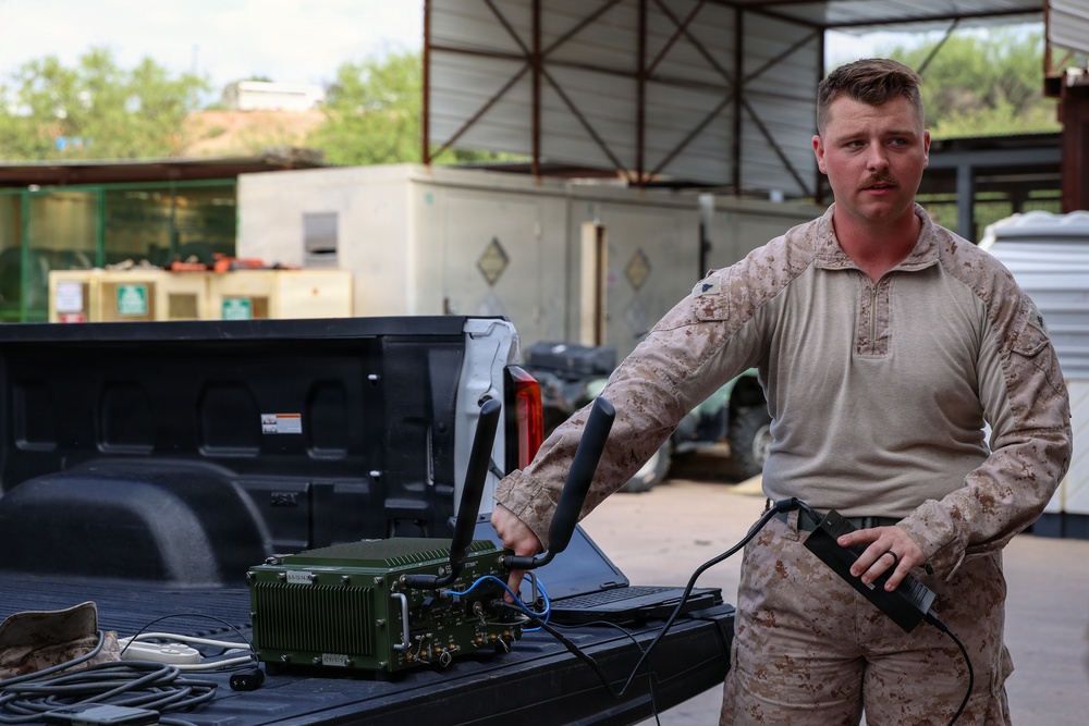 A U.S. Marine in desert MARPAT camouflage uniform is standing at the bed of a pickup truck, handling military communications equipment. He appears to be demonstrating or setting up a green tactical radio/communications unit with antennas, while holding a handheld device in his other hand. The setting appears to be a forward operating base or military installation with vehicles and structures visible in the background.