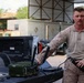 A U.S. Marine in desert MARPAT camouflage uniform is standing at the bed of a pickup truck, handling military communications equipment. He appears to be demonstrating or setting up a green tactical radio/communications unit with antennas, while holding a handheld device in his other hand. The setting appears to be a forward operating base or military installation with vehicles and structures visible in the background.