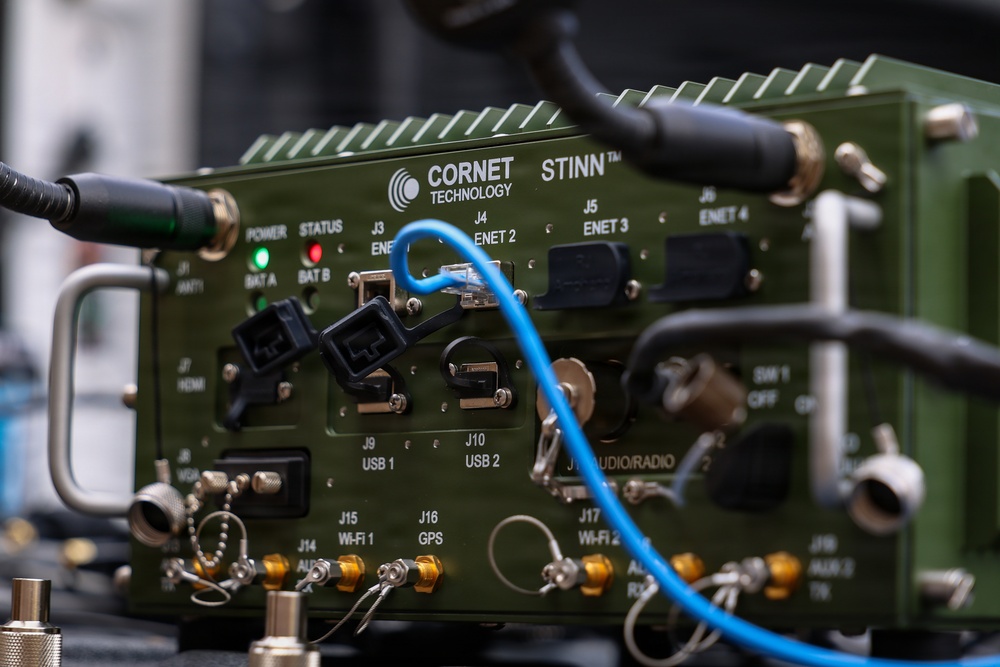 The image shows a military-grade electronic communications device manufactured by Cornet Technology, model STINN™, housed in an olive-drab green ruggedized enclosure. The unit features multiple labeled ports and connectors including Ethernet, USB, GPS, Wi-Fi, HDMI, and audio/radio interfaces. Several cables are actively connected to the device, including a visible blue Ethernet cable.