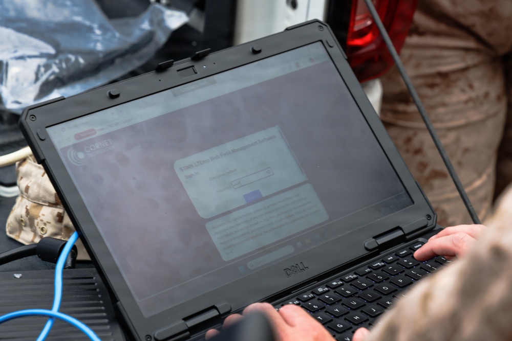 A person in military camouflage uniform is operating a ruggedized Dell laptop in what appears to be a field environment. The laptop screen displays a CORNET-branded web interface showing a 'STNN LTEmin Web Field Management Software' sign-in page. The setup includes a blue ethernet/data cable connected to the laptop, suggesting field communications or network management operations.