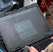 A person in military camouflage uniform is operating a ruggedized Dell laptop in what appears to be a field environment. The laptop screen displays a CORNET-branded web interface showing a 'STNN LTEmin Web Field Management Software' sign-in page. The setup includes a blue ethernet/data cable connected to the laptop, suggesting field communications or network management operations.