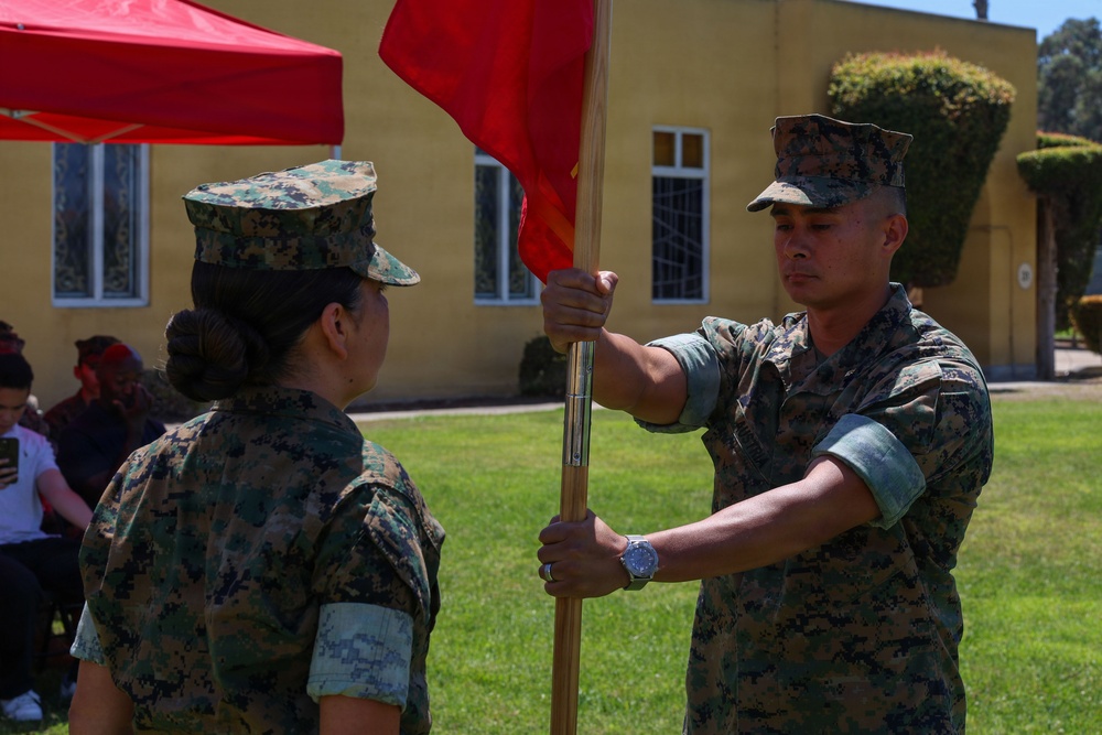 Two U.S. Marines in MARPAT camouflage uniforms are participating in a formal guidon transfer ceremony outdoors. One Marine is passing a red guidon (unit flag) on a wooden staff to the other, a ritual commonly associated with a change of command or responsibility. The ceremony appears to be taking place on a military installation with an audience seated in the background.