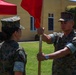 Two U.S. Marines in MARPAT camouflage uniforms are participating in a formal guidon transfer ceremony outdoors. One Marine is passing a red guidon (unit flag) on a wooden staff to the other, a ritual commonly associated with a change of command or responsibility. The ceremony appears to be taking place on a military installation with an audience seated in the background.
