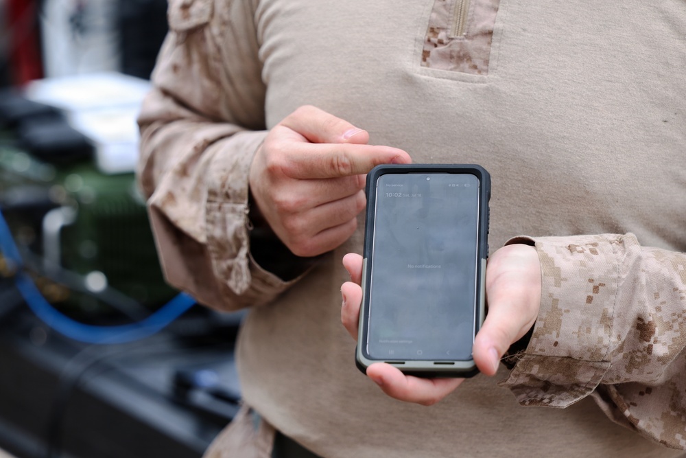 A person wearing U.S. Marine Corps MARPAT desert camouflage uniform is holding and interacting with a smartphone in a rugged protective case. The phone's lock screen is visible, displaying a time of 10:02. The setting appears to be an outdoor or semi-industrial military environment.