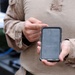A person wearing U.S. Marine Corps MARPAT desert camouflage uniform is holding and interacting with a smartphone in a rugged protective case. The phone's lock screen is visible, displaying a time of 10:02. The setting appears to be an outdoor or semi-industrial military environment.