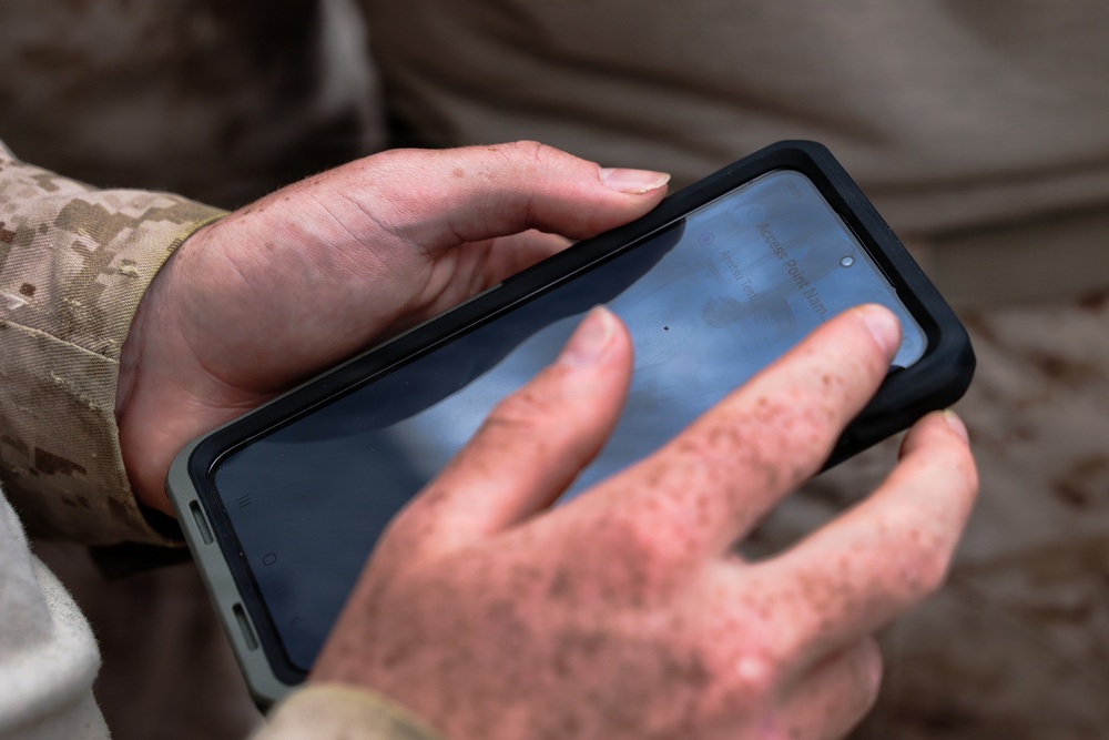 A person wearing desert camouflage military uniform is holding a smartphone in a black protective case. The phone screen is partially visible and appears to display text including what looks like 'Access Point Name'. The hands are visibly dirty or mud-stained, suggesting an outdoor field environment.