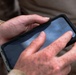 A person wearing desert camouflage military uniform is holding a smartphone in a black protective case. The phone screen is partially visible and appears to display text including what looks like 'Access Point Name'. The hands are visibly dirty or mud-stained, suggesting an outdoor field environment.