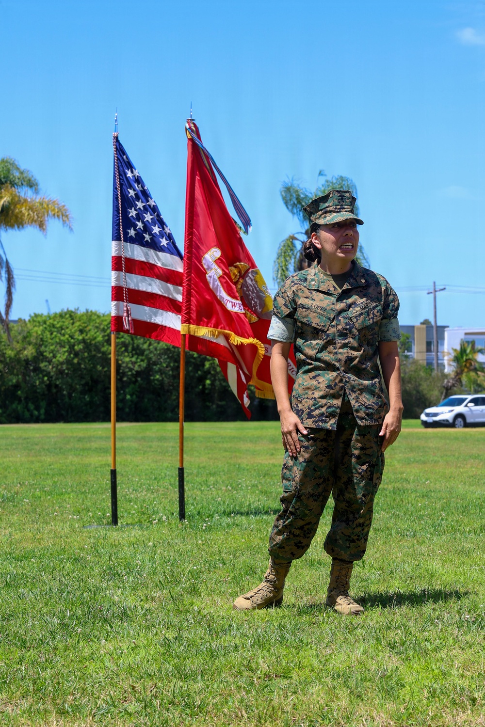 A female U.S. Marine in MARPAT woodland camouflage utility uniform stands at attention on a grass field during what appears to be a military ceremony. She is positioned in front of the American flag and a red U.S. Marine Corps unit guidon/colors. The setting appears to be a military installation, likely on the West Coast based on the palm trees and vegetation.