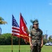 A female U.S. Marine in MARPAT woodland camouflage utility uniform stands at attention on a grass field during what appears to be a military ceremony. She is positioned in front of the American flag and a red U.S. Marine Corps unit guidon/colors. The setting appears to be a military installation, likely on the West Coast based on the palm trees and vegetation.