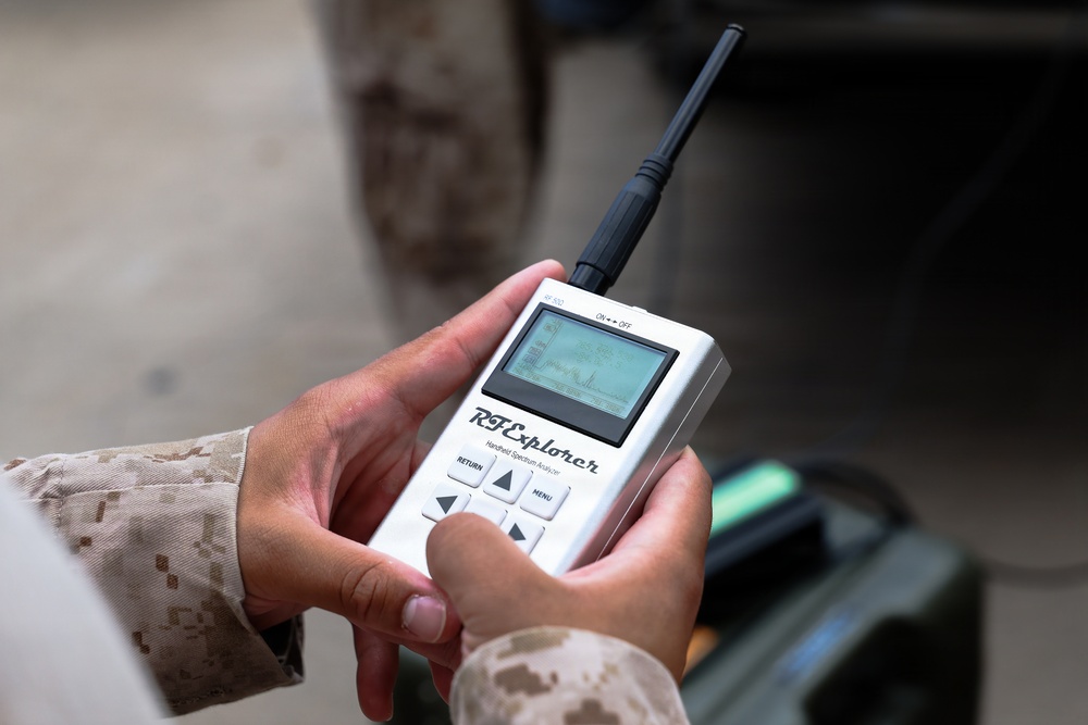 A person wearing military desert camouflage (MARPAT pattern) is holding and operating an RF Explorer handheld spectrum analyzer. The device is white, has a small LCD screen displaying a frequency spectrum graph, a black antenna, and navigation buttons. The background is blurred but suggests an outdoor or semi-covered military environment.