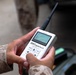 A person wearing military desert camouflage (MARPAT pattern) is holding and operating an RF Explorer handheld spectrum analyzer. The device is white, has a small LCD screen displaying a frequency spectrum graph, a black antenna, and navigation buttons. The background is blurred but suggests an outdoor or semi-covered military environment.