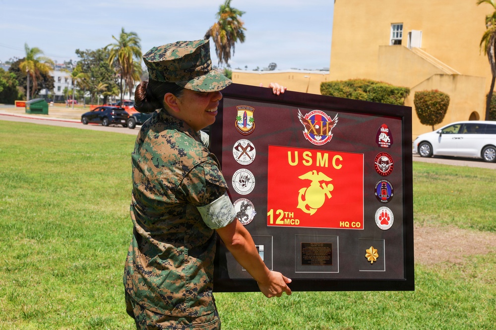 A female U.S. Marine in MARPAT digital camouflage uniform is holding a large commemorative shadow box/plaque outdoors on a military installation. The framed display prominently features USMC insignia, the text '12th MCD HQ CO', and multiple unit patches and coins. The setting appears to be a well-maintained base grounds with palm trees and tan military buildings visible in the background.