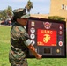 A female U.S. Marine in MARPAT digital camouflage uniform is holding a large commemorative shadow box/plaque outdoors on a military installation. The framed display prominently features USMC insignia, the text '12th MCD HQ CO', and multiple unit patches and coins. The setting appears to be a well-maintained base grounds with palm trees and tan military buildings visible in the background.