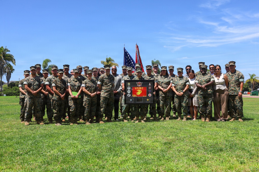 A large group of U.S. Marine Corps personnel and several civilians pose for a formal group photograph outdoors on a grassy field. The group is centered around a framed USMC commemorative plaque or shadow box being held by individuals in the front center. Two flags — the American flag and the USMC flag — are visible in the background.