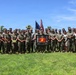 A large group of U.S. Marine Corps personnel and several civilians pose for a formal group photograph outdoors on a grassy field. The group is centered around a framed USMC commemorative plaque or shadow box being held by individuals in the front center. Two flags — the American flag and the USMC flag — are visible in the background.