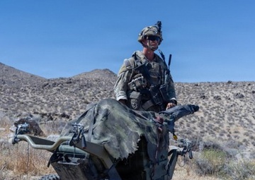 A soldier in full combat gear and camouflage uniform is seated on a small military quad-bike or all-terrain vehicle (ATV) in an arid desert environment. The vehicle appears to be a compact, military-spec four-wheeled unit covered with a camouflage net or drape. The setting suggests a training exercise or field operation in a desert region.