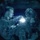LCF-24: Lowlight Fast-roping drills Two military personnel are photographed in low-light conditions, likely inside a vehicle or enclosed space. One individual is using a small bright light source, possibly a flashlight or penlight, directed toward the other person's head area. The image has a distinctive circular vignette framing, suggesting it may have been captured through night-vision or a specialized lens.
