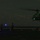 LCF-24: Lowlight Fast-roping drills A nighttime helicopter landing or departure operation on what appears to be a naval vessel flight deck. Two deck crew members are visible using illuminated wands/batons to guide the helicopter, which is displaying green navigation lights. The scene is very dark with only artificial lighting sources visible.