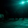 LCF-24: Lowlight Fast-roping drills A nighttime military operation on what appears to be a naval vessel flight deck, illuminated by intense green-tinted overhead lighting. Several personnel in tactical gear and helmets are visible standing near a helicopter, which is chained to the deck. The wet reflective surface and dark surroundings suggest an at-sea environment during night operations.