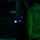 LCF-24: Lowlight Fast-roping drills A person wearing a high-visibility safety vest marked 'LPD-17' is photographed from behind at night. They are holding a device emitting two bright white lights, likely a flashlight or signaling device. The scene is illuminated with a green ambient light, consistent with a naval or military flight deck environment.