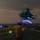 LCF-24: Lowlight Fast-roping drills A night operations scene on a naval vessel's flight deck showing a deck handler guiding a military helicopter during landing or takeoff operations. The handler is wearing a yellow jersey and helmet, using illuminated wands to signal the aircraft. The helicopter, likely a Bell AH-1Z Viper or similar attack/utility helicopter, is hovering with green navigation lights and cockpit illumination visible.