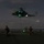 LCF-24: Lowlight Fast-roping drills A nighttime military operation on what appears to be a naval vessel's flight deck, with a helicopter hovering overhead connected by a rope or cable to the deck below. Three armed military personnel in full combat gear are positioned in kneeling and standing stances on the flight deck. The helicopter displays green NVIS (Night Vision Imaging System) compatible lighting, indicating a covert or low-visibility operation.