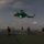 LCF-24: Lowlight Fast-roping drills A military helicopter hovers over a naval vessel's flight deck at dusk or night, with green infrared/NVIS lighting visible on the aircraft. Several armed personnel in full combat gear are positioned on the deck in tactical stances, and a rope or fast-rope line descends from the helicopter toward the deck. This appears to be a fast-rope insertion/extraction (FRIES) or similar special operations training exercise conducted at night.