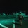 LCF-24: Lowlight Fast-roping drills A nighttime military operation on what appears to be a naval vessel flight deck, illuminated by green-tinted lighting. Multiple armed and equipped soldiers in full combat gear are moving across the deck toward helicopters. At least two military helicopters are visible on the deck, with additional personnel in colored helmets stationed nearby.