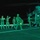 LCF-24: Lowlight Fast-roping drills A nighttime military operation showing armed personnel boarding or preparing to board a helicopter marked with the number '67' on what appears to be a ship's flight deck or airfield. The scene is illuminated by green lighting, consistent with night operations or green-filtered lighting used to preserve night vision. Multiple soldiers in full combat gear are moving in formation toward the aircraft.