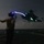 LCF-24: Lowlight Fast-roping drills A naval flight deck crew member is directing a helicopter during a nighttime operation aboard a warship. The handler is using illuminated wands/batons to signal the aircraft, which appears to be lifting off or hovering near the flight deck. The long-exposure photograph captures light trails from the wands, creating streaks of blue and white light against the dark sky.