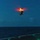 LCF-24: Lowlight Fast-roping drills A military or coast guard helicopter is captured in flight at dusk or nighttime over open ocean, photographed from the deck of a naval vessel. The rotor blades are illuminated with a bright orange-red glow, likely from rotor tip lights or a long-exposure camera effect. The ship's flight deck is partially visible in the lower right corner with green deck lighting.