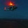 LCF-24: Lowlight Fast-roping drills A military helicopter is captured hovering or taking off at dusk/night over what appears to be a ship's flight deck or airfield. The rotor blades are illuminated by an orange-red anti-collision beacon, creating a dramatic light trail effect. Navigation lights (green and red) are visible on the aircraft against the deep blue twilight sky.