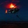 LCF-24: Lowlight Fast-roping drills A military helicopter is captured mid-flight at night, likely lifting off from or hovering over a ship's flight deck. The rotor blades create a dramatic glowing orange disc effect due to a rotor tip light and long exposure photography. Navigation lights in red and green are visible on the aircraft, and blue deck lighting marks the landing area below.