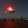 LCF-24: Lowlight Fast-roping drills A military helicopter is hovering or landing on a naval vessel's flight deck at night, illuminated by red and green navigation lights. Several personnel in full tactical gear are positioned on the deck around the aircraft. The scene appears to depict a nighttime helicopter operations exercise or insertion/extraction mission aboard a warship.