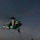 LCF-24: Lowlight Fast-roping drills A military helicopter, appearing to be a UH-1Y Venom or AH-1Z Viper type, is captured lifting off or hovering low over a naval vessel's flight deck at night. The aircraft displays bright green navigation/IR lights and bears a visible side number. A single deck crew member in flight gear stands on the darkened flight deck observing the aircraft.